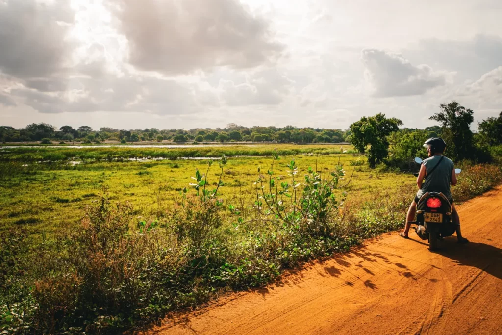 A man sitting on a motorbike on a dirt track looking over farmland and lakes near Arugam Bay