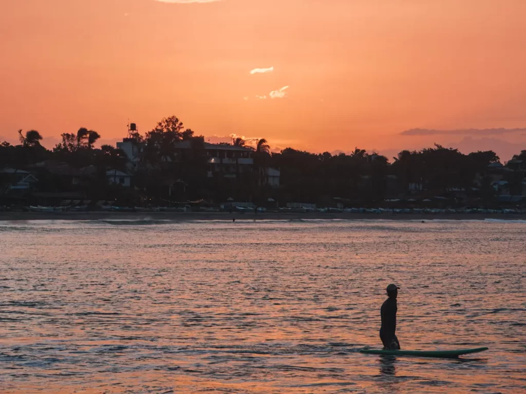 A surfer walks with his board witht he sun setting in the background
