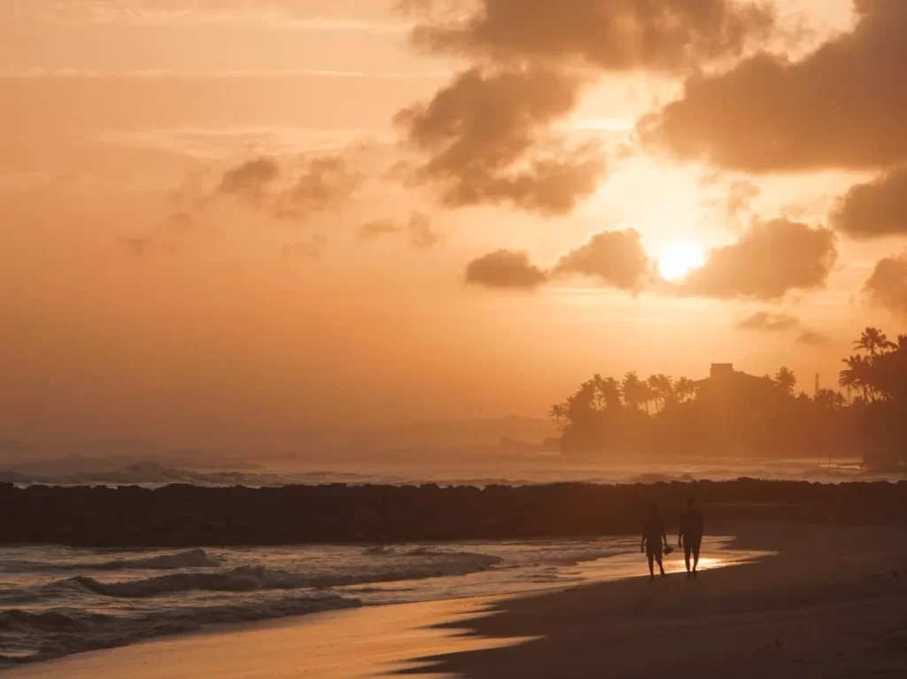 Two people walk down an empty beach with a bright orange sky and the sunsetting behind the headlands in Ahangama
