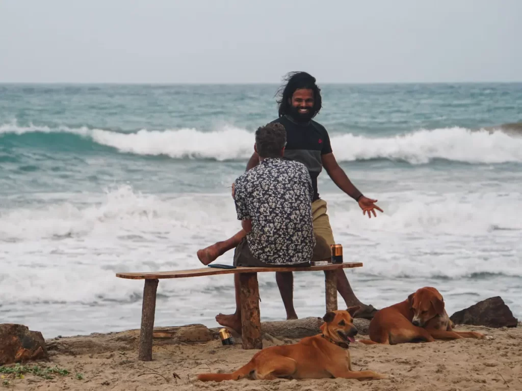 Two guys chat on a bench with dogs around them and the ocean in the background